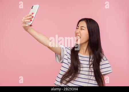 Portrait de fille asiatique fille puérile avec des cheveux brunette prenant selfie et démontrant la langue, faisant le visage s'amuser tout en faisant la photo pour le blog. Banque D'Images