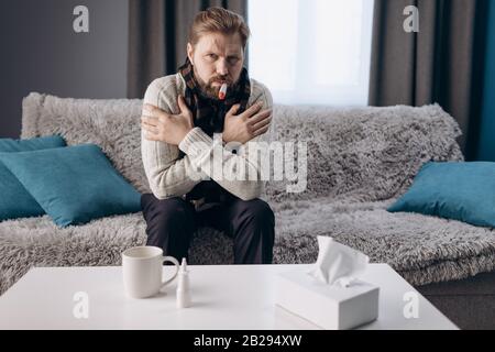 Homme fatigué barbu dans des vêtements chauds et écharpe tricotée mesurant la température du corps avec thermomètre tout en étant assis sur un canapé gris. Personne d'âge mûr prenant la voiture Banque D'Images