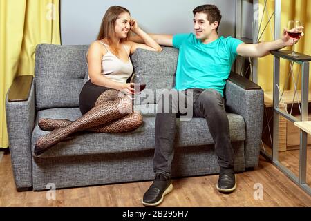 Une date d'un jeune homme et d'une femme à l'intérieur, les amoureux sont assis sur le canapé avec des verres de vin dans leurs mains. Réunion romantique avec boissons alcoolisées. Banque D'Images