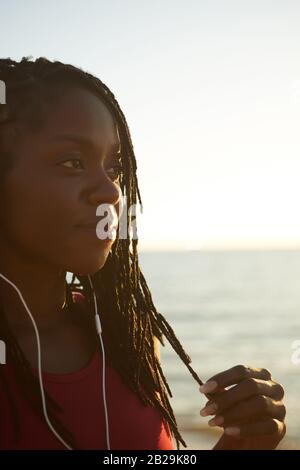 Jeune femme noire souriante pensive qui touche ses dreadlocks et regarde loin lors de la marche sur la plage Banque D'Images