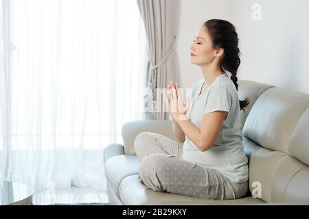 Belle jeune femme enceinte méditant en position lotus lorsqu'elle est assise sur un canapé confortable à la maison Banque D'Images