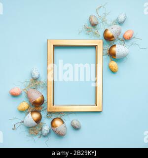 Vue sur le dessus des œufs de Pâques colorés dans différentes couleurs disposés autour d'un cadre photo doré. Fond bleu. Espace de copie. Banque D'Images