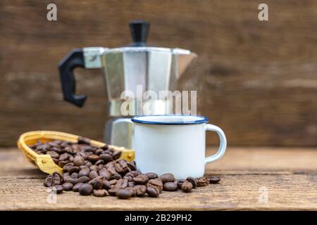 Une tasse de café chaud en étain blanc avec une cafetière italienne (Moka) et des grains de café sur une table en bois. Banque D'Images