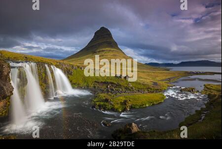 Montagne Kirkjufell ensoleillée contre les nuages gris fond. Cascades avec arc-en-ciel en premier plan. Banque D'Images