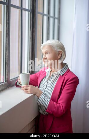 Une dame souriante aux cheveux gris se tenant à la fenêtre et pensive Banque D'Images