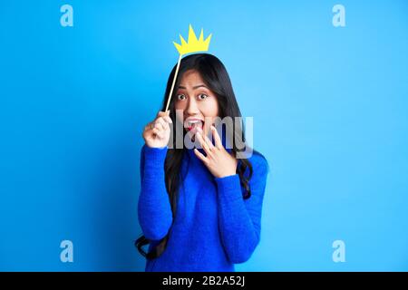 Une belle femme surprise tient une couronne d'artisanat en papier sur un bâton sur fond bleu. Parti, concept d'émotions Banque D'Images