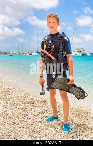 Plongeur masculin néerlandais posant sur la plage de Bonaire avec l'océan et les bateaux Banque D'Images