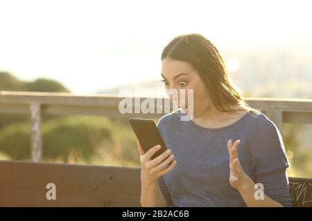 Surprise femme regardant choqué son écran de smartphone sur un banc de parc au coucher du soleil Banque D'Images