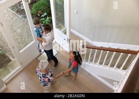 Vue en grand angle d'un jeune frère caucasien et sœur à la maison, ouvrant la porte d'entrée et accueillant leurs grands-parents, grand-père transportant sa gra Banque D'Images