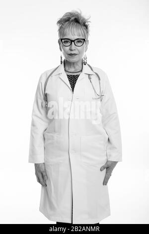 Studio shot of senior Asian woman doctor standing tout en portant des lunettes contre fond blanc Banque D'Images