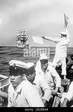 Vue sur la mer au large de la côte japonaise. Au premier plan se trouvent certains cadets de l'École navale de Tokyo de la Marine impériale japonaise dans un bateau à ramer. L'un d'entre eux est en contact avec son navire de formation, le Taisei Maru, au moyen de drapeaux de signalisation. Banque D'Images