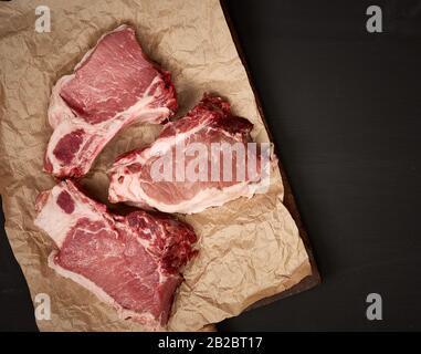 tranches de porc juteux cru de viande sur la côte, la nourriture se trouve sur du papier parchemin brun, fond en bois noir, vue de dessus Banque D'Images