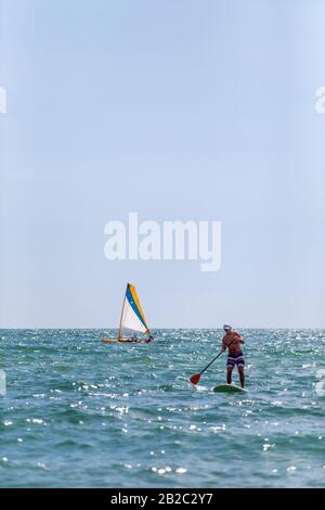 Homme qui utilise sa palette sur un panneau de paddle debout avec un voilier en arrière-plan dans le golfe du Mexique près de Naples, en Floride Banque D'Images