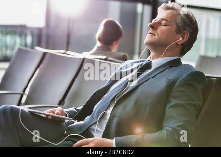 Homme d'affaires mûr dormant tout en écoutant de la musique avec des écouteurs à l'aéroport avec une lumière diffuse Banque D'Images