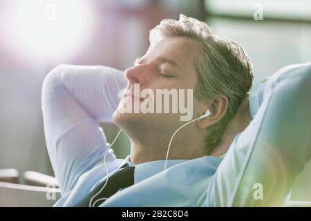 Homme d'affaires mûr dormant tout en écoutant de la musique avec des écouteurs à l'aéroport avec une lumière diffuse Banque D'Images