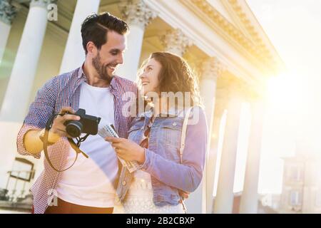 Portrait de beau jeune homme montrant des photos sur l'appareil photo à sa belle petite amie tout en étant assis contre le musée avec l'éclat de lentille en arrière-plan Banque D'Images