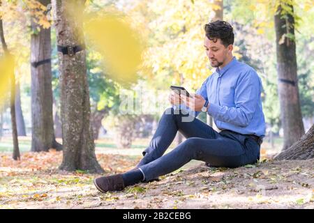 Beau jeune homme lisant un ebook dans un parc. Portrait d'un jeune homme avec une veste denim et une chemise bleue lisant un e-livre à l'extérieur. Un gars qui lit un Banque D'Images