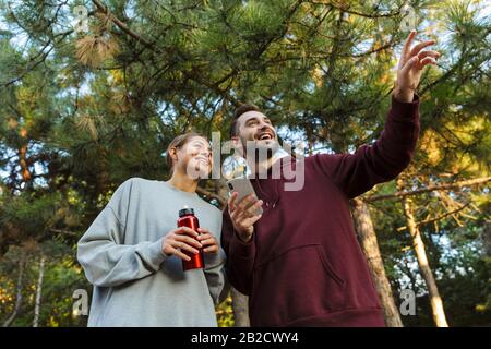 Photo d'une jeune heureuse heureuse femme de sport positive et homme utilisant le téléphone mobile à l'extérieur dans le parc vert de la nature pointant de côté tenant bouteille avec wat Banque D'Images