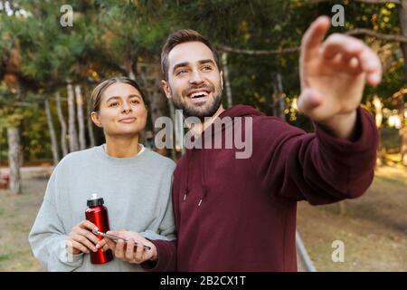 Photo d'une heureuse femme de sport positive et l'homme utilisant le téléphone mobile à l'extérieur dans la nature parc vert pointant de côté tenant bouteille avec de l'eau. Banque D'Images