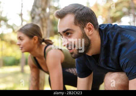 Photo de jeune couple heureux dans les vêtements de sport faire des exercices tout en s'exerçant dans le parc vert pendant la journée ensoleillée d'été Banque D'Images