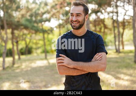 Image d'un jeune homme sportif fort et optimiste et gai qui se pose dehors dans la nature parc vert regardant de côté. Banque D'Images