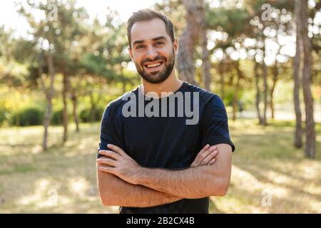 Image d'un jeune homme de sport heureux et heureux se posant dehors dans la nature parc vert regardant caméra. Banque D'Images