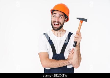 Photo de jeune homme optimiste constructeur dans casque posant isolé sur fond de mur blanc tenant marteau. Banque D'Images