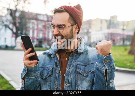 Gros plan sur un heureux jeune homme barbu élégant marchant dehors dans la rue, en utilisant le téléphone mobile, célébrant le succès Banque D'Images