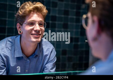 Jeune homme avec des lunettes et un sourire perçant devant un miroir où il se voit se refléter dans une salle de bains Banque D'Images