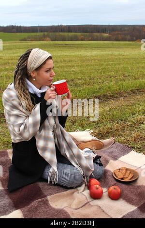 Syktyvkar, Russie - septembre 2019: Une belle fille d'apparence européenne avec de longs verrous boit du thé et semble pensivement dans la distance. Pique-nique dans la nature. Photographie d'automne. Banque D'Images