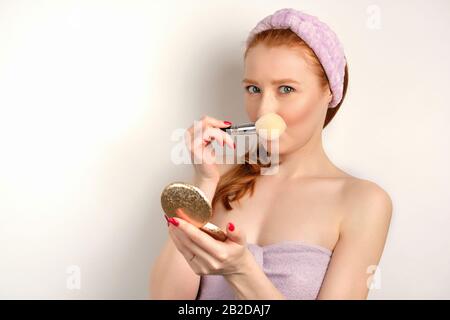 Une fille à poil rouge dans une serviette lilas se tient sur un fond blanc, recouverte d'une brosse et s'insinant dans le cadre Banque D'Images