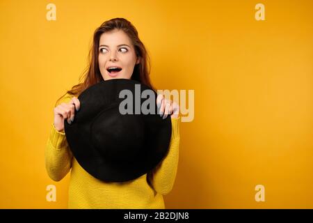 Belle brunette dans un chandail jaune regarde avec enthousiasme, se cachant derrière un chapeau noir sur un fond jaune. Banque D'Images