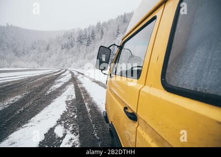 Carpathians, Ukraine - décembre 2019: Camionnette de camping-cars jaune conduite sur la route enneigée de terre avec forêt d'hiver sur fond. Banque D'Images