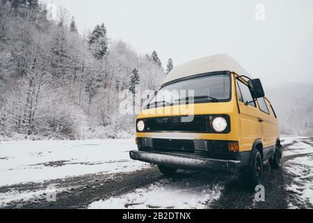 Carpathians, Ukraine - décembre 2019: Camionnette de camping-cars jaune conduite sur la route enneigée de terre avec forêt d'hiver sur fond. Banque D'Images