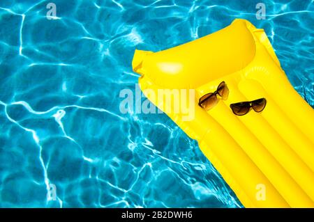 Ses lunettes de soleil et ses hers reposent sur un radeau gonflable jaune vide flottant dans une piscine bleu vif Banque D'Images