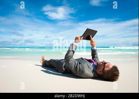 Homme d'affaires détendu allongé dans un costume à l'aide de son ordinateur tablette sur le rivage d'une plage tropicale Banque D'Images