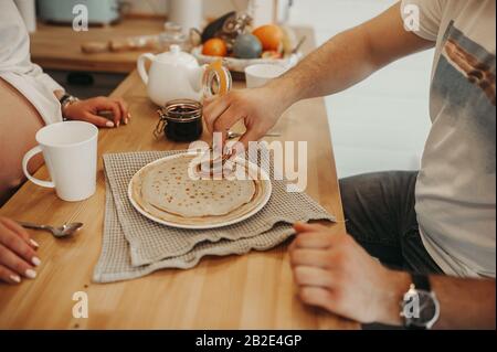 une paire d'amoureux avec une fille enceinte assise à une table en bois, boire du thé, manger des crêpes avec confiture dans une cuisine confortable. Crêpes chaudes sur un pla blanc Banque D'Images