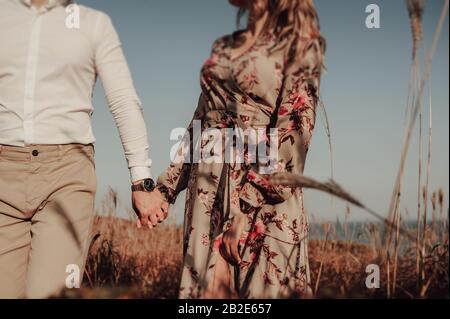 deux amoureux, une femme dans une robe à imprimé et un homme dans une chemise blanche tenant les mains au milieu d'un champ de blé en automne Banque D'Images