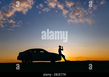 Silhouette d'homme conducteur se reposant après une promenade, assis sur la capuche de sa voiture et de l'eau potable d'une bouteille, vue latérale. Heure du coucher du soleil. Banque D'Images