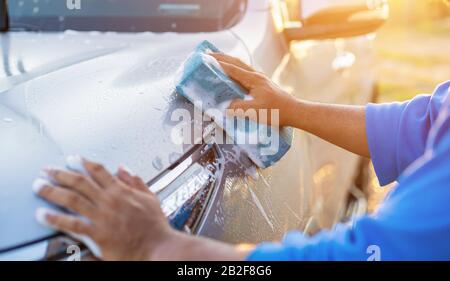 Homme asiatique utilisant une éponge bleue avec du savon pour laver la voiture à l'extérieur au coucher du soleil. Concept de nettoyage et d'entretien de voiture Banque D'Images
