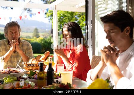 Vue de face d'un homme caucasien et de sa fille et de son fils adultes assis dehors à une table de dîner pour un repas, avec des mains claquées et des yeux fermés dans p Banque D'Images