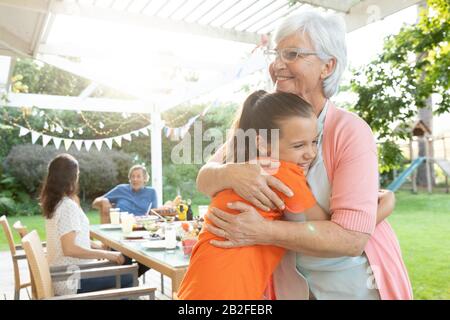 Vue latérale d'une femme caucasienne senior et de sa jeune petite-fille debout et embrassant dehors dans un jardin, avec leur famille assis à un dîner ta Banque D'Images