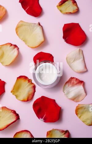 Hydratant ouvert sur un fond rose pastel parmi les pétales de rose rouge et orange. Le concept des soins de la peau, des soins anti-vieillissement, des ingrédients naturels dans les cosmétiques Banque D'Images