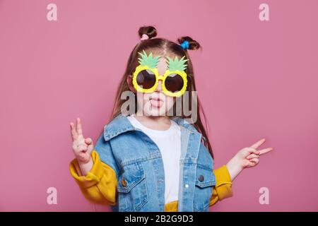 Concept de mode d'été. Petite fille gaie dans de grandes lunettes de soleil ananas sur fond rose Banque D'Images