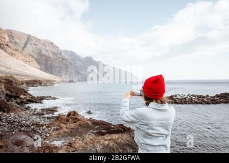 Jeune femme à Red Hat en profitant d'un voyage sur une côte rocheuse de l'océan, en photographiant au téléphone des paysages à couper le souffle. Voyager sur l'île de Tenerife, Espagne Banque D'Images