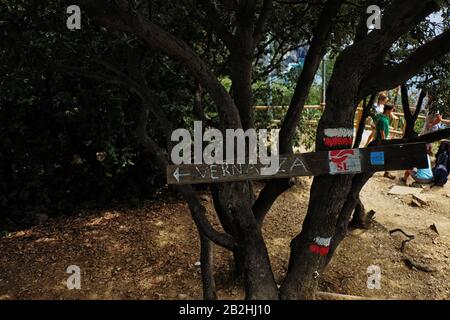 Un signe d'une planche clouée à un arbre marquant le chemin vers Vernazza, arbustes et personnes reposant sur le chemin en arrière-plan Banque D'Images