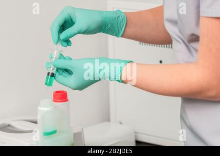 Les mains dans des gants verts déverse le liquide vert de la seringue dans un récipient en plastique. Banque D'Images