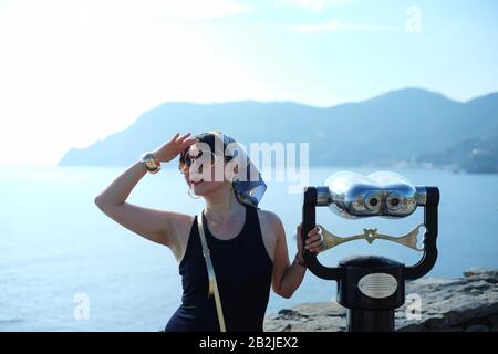 Vernazza, Cinque Terre une femme glamour en foulard et lunettes de soleil tenant une grande jumelle à un belvédère vue sur la baie et les montagnes de Monterosso Banque D'Images