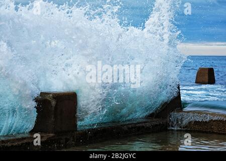 Brisant les vagues, l'eau gelée dans le temps comme une sculpture de verre contre les murs de périmètre en béton crénelés du Ross Jones Rockpool Coogee, Sydney Banque D'Images