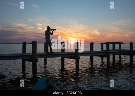 Photographe solitaire se tenant sur la jetée de Chesapeake Bay, silhouetted contre un coucher de soleil bleu et or Banque D'Images